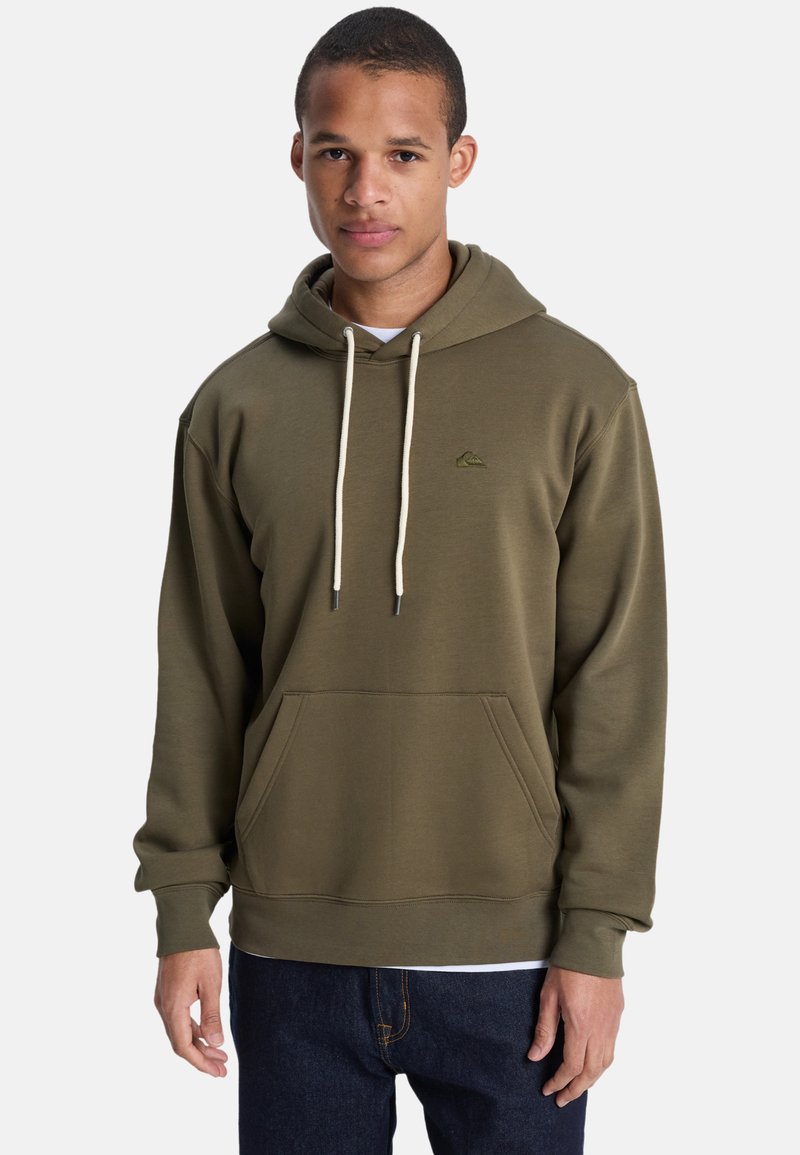 Jeune homme portant un sweat à capuche vert olive avec des cordons blancs et un jean bleu foncé, debout devant un fond clair uni.