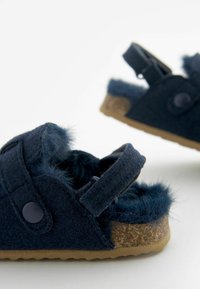 Chaussons en feutre bleu marine avec accents en fourrure, bande réglable et semelle en liège. Présente un détail de bouton rond. Surface texturée pour le confort.