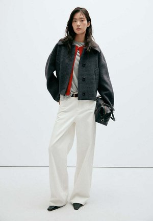 Mujer con chaqueta negra recortada, cárdigan rojo, top gris, pantalones blancos de pierna ancha y zapatos negros puntiagudos, sosteniendo un bolso negro sobre un fondo blanco.