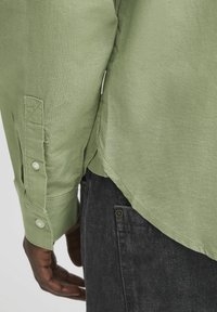 Camicia in lino verde chiaro con maniche lunghe, polsini abbottonati e orlo curvo, abbinata a jeans in denim grigio scuro.