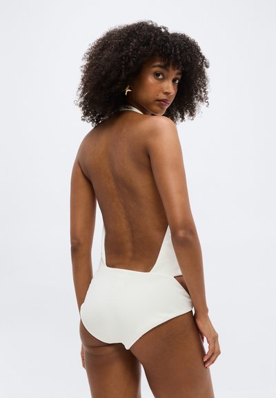 Kvinde med krøllet hår iført en hvid rygfri badedragt, der kigger over sin skulder mod en ensartet hvid baggrund.