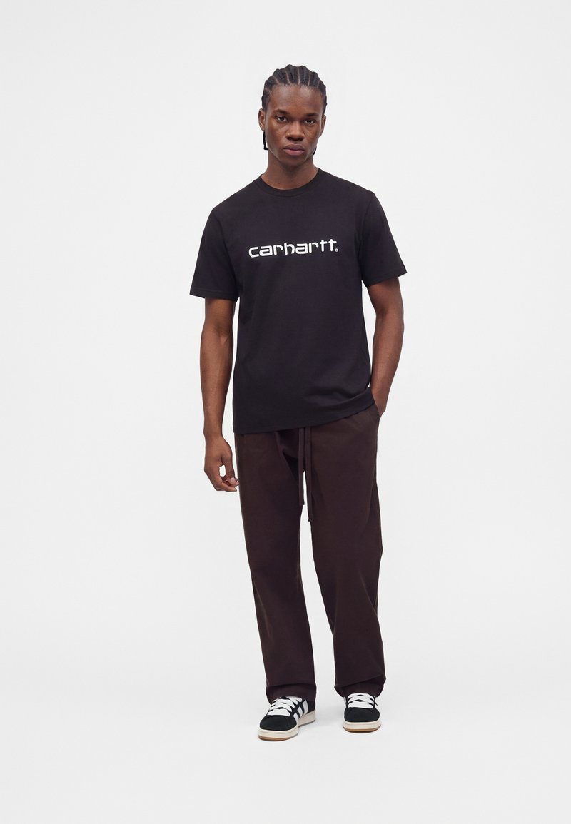 Junger Mann mit geflochtenem Haar, der ein schwarzes Carhartt-T-Shirt, dunkelbraune Hose und schwarz-weiße Sneaker vor einem einfarbigen Hintergrund trägt.