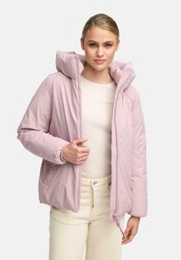 Vrouw in een lichtroze gewatteerde jas met capuchon over een crèmekleurige geribde trui en crèmekleurige hoge taille broek, staand tegen een effen achtergrond.