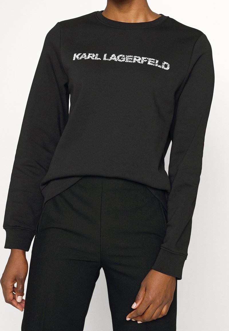 Femme portant un haut noir à manches longues avec le texte "KARL LAGERFELD" sur la poitrine et un pantalon noir assorti sur fond blanc.