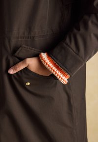 Veste imperméable marron avec coutures apparentes, une poche latérale et un détail de poignet en crochet orange et blanc. Un bouton-pression doré est présent.