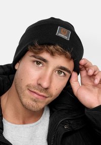 Zwarte gebreide beanie met een klein rechthoekig logo-patch met "JEANSCODE" in grijs en oranje. Gladde textuur, stevige pasvorm en ronde top.