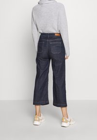 Dark blue wide-leg jeans with a high waist, back pockets, and stitching details, paired with light-colored sneakers. Grey sweater visible.