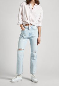 Woman wearing light blue ripped jeans, white sneakers, and a white long-sleeve shirt tied at the waist, standing with one hand in pocket.