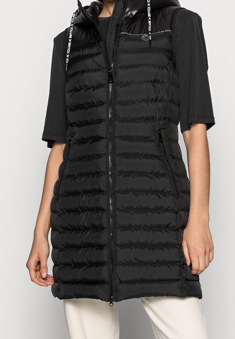 Czarna pikowana kamizelka z zamkiem błyskawicznym z przodu, kapturem i bocznymi kieszeniami. Charakteryzuje się pikowaną teksturą oraz odblaskowymi troczkami do zapięcia.