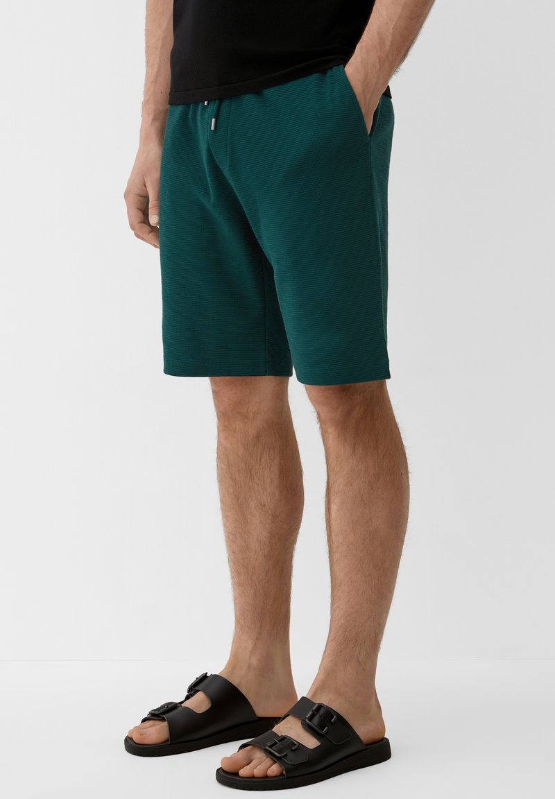 Homme portant un short bleu sarcelle jusqu'aux genoux avec des poches latérales, un haut noir et des sandales noires à orteils ouverts avec des brides à double boucle, debout sur un sol blanc.