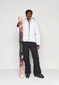 Person in weißer Skijacke und schwarzen Hosen hält pinke Skier senkrecht, trägt Skischuhe und schwarze Skibrille auf dem Kopf.