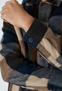 Ruud flanellsärk sinise, pruuni ja beeži varjundites; sellel on must nupukinnitus ja tekstuuriga kangas kontrastsete varrukatega.