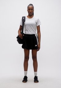 Γυναίκα όρθια που φοράει λευκό μπλουζάκι Adidas, μαύρη φούστα Adidas, μαύρα αθλητικά παπούτσια, κάλτσες και κρατάει μια μαύρη αθλητική τσάντα πάνω σε έναν ώμο.