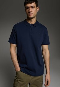 Granatowa koszulka polo wykonana z bawełny, z klasycznym kołnierzykiem, zapięciem na dwa guziki, krótkimi rękawami oraz małym logo na lewej piersi.