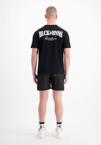 Zwarte katoenen t-shirt met witte bedrukking op de achterkant, gecombineerd met donkere shorts. De schoenen van het model zijn wit, met zwarte en gele accenten.