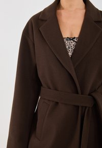 Donna che indossa un cappotto brown scuro con cintura sopra una maglietta a stampa leopardata con un piccolo fiocco dettaglio sul collo.