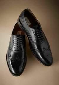 Zwarte leren brogues met decoratieve perforaties, veters en lage hakken op een beige achtergrond.