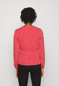 Blusa rossa con un motivo a pois bianchi, maniche lunghe e vita aderente. Indossata con pantaloni neri, su uno sfondo semplice.