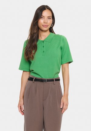 Groene polo met korte mouwen, voorzien van een gestructureerde brei, met een knoopsluiting bestaande uit drie knopen, gedragen met bruin high-waisted broeken en een zwarte riem.