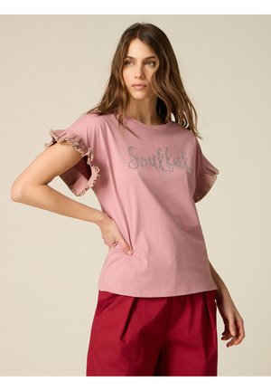 Ung kvinde med langt brunt hår iført en lyserød t-shirt med flæser på ærmerne med teksten "Soulful" og røde bukser, poserende med den ene hånd på hoften.