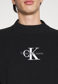 Schwarzer gerippter Strickpullover mit hohem Kragen und einem weißen gestickten Logo, das "Calvin Klein Jeans" über die Brust trägt.