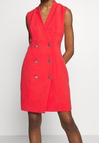 Robe sans manches rouge croisée à double boutonnage, longueur genou, avec des boutons argentés, portée par une personne debout sur un fond uni.