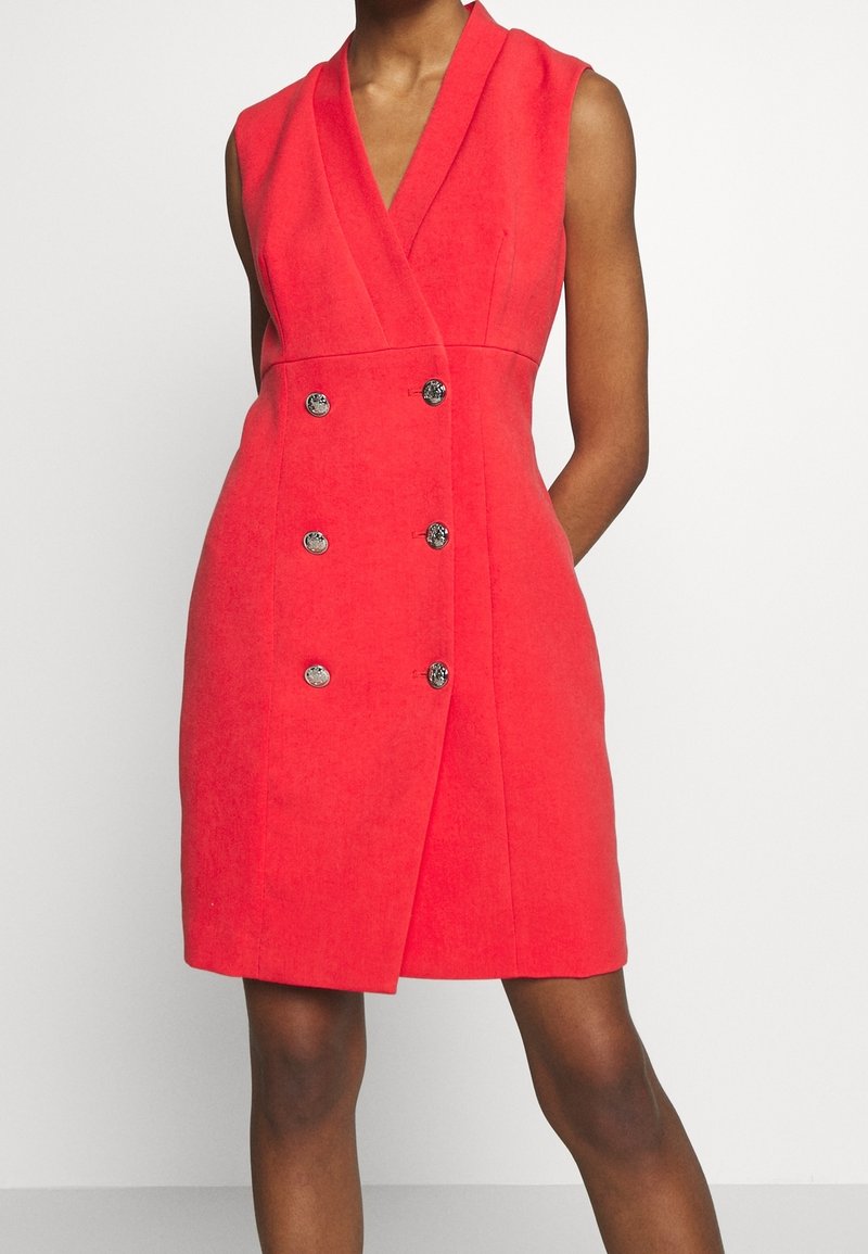 Robe sans manches rouge croisée à double boutonnage, longueur genou, avec des boutons argentés, portée par une personne debout sur un fond uni.