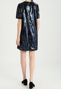 Kvinna som står, klädd i en kortärmad klänning med svarta och blå paljetter samt svarta klackskor, vänd bort från kameran mot en enfärgad bakgrund.