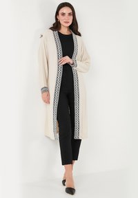Beige cardigan with black lace trim, open front, long sleeves, designed with a relaxed fit. Worn over a black outfit with fitted pants.