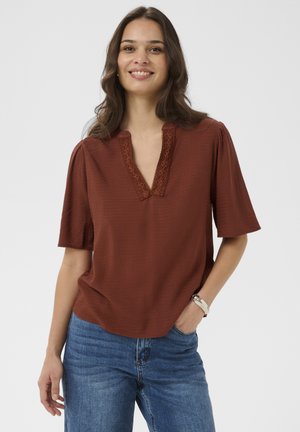 Smilende kvinde med bølget brunt hår iført en rustfarvet bluse med blonder ved halsudskæringen og blå jeans, hænder afslappet ved siden.