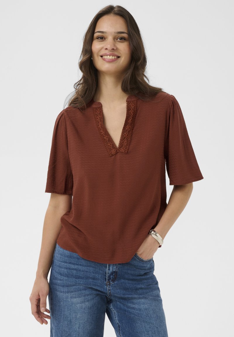 Femme souriante aux cheveux bruns ondulés portant un chemisier couleur rouille avec un col en dentelle et un jean bleu, les mains détendues le long du corps.