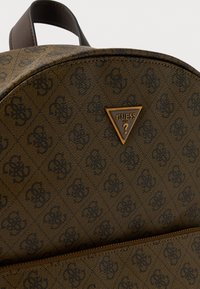 Mochila marrón con patrón y superficie texturizada, que presenta un logo triangular dorado y detalles en cuero marrón. Compartimento con cremallera visible.