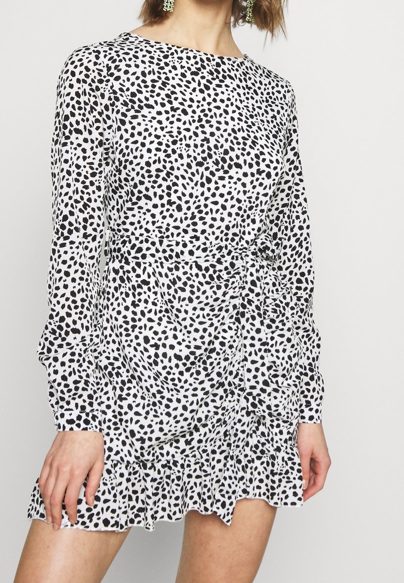 Femme portant une robe blanche courte à manches longues avec des pois noirs irréguliers et un ourlet à volants, debout devant un fond uni.