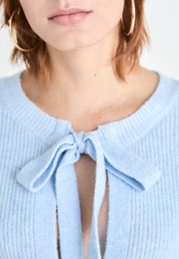 Pull bleu clair en maille avec un col rond et un détail noué à l'avant, présentant une texture douce et un motif côtelé.