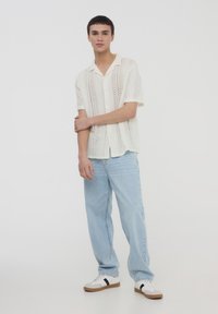 Camicetta a maniche corte di colore crema con un design in pizzo, abbinata a jeans a gamba dritta blu chiaro e sneakers bianche con accenti neri.