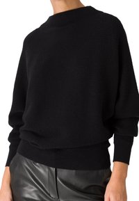Personne portant un pull en maille côtelée noir à manches longues associé à un pantalon en cuir noir, raccourci à mi-torse.
