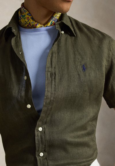 Άνδρας που φοράει ένα πουκάμισο σε ελαιί πράσινο με κουμπιά πάνω από ένα ανοιχτό μπλε T-shirt και ένα κίτρινο φλοράλ μαντήλι στον λαιμό, εμφανιζόμενος από τους ώμους μέχρι τη μέση.