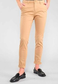 Person, der beige schmal geschnittene Hosen mit hochgerollten Säumen und glänzenden schwarzen Slippern trägt, steht mit Händen in den Taschen vor einem schlichten Hintergrund.