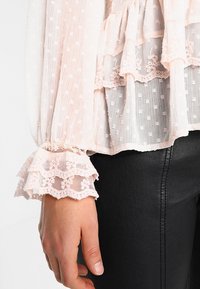 Jasnoróżowa bluzka z długim rękawem, o teksturowanej tkaninie, z warstwowymi falbanami i koronkowymi wykończeniami przy mankietach, zestawiona z czarnymi skórzanymi spodniami.