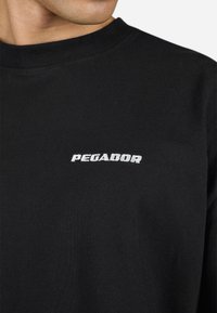 Chemise noire à manches longues en coton doux. Présente un logo blanc "PEGADOR" sur la poitrine, avec un design classique à col rond.