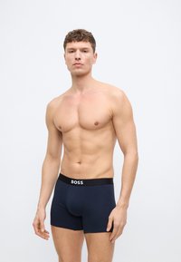 Μυώδης άνδρας όρθιος, φοράει στενά μπλε σκούρα μποξεράκια με μαύρη ζώνη μέσης με την ένδειξη "BOSS", μπροστά σε απλό φωτεινό φόντο.