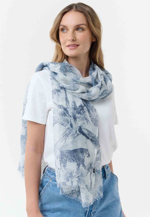 Ženska, oblečena v svetlo modro ruto z živalskim in rastlinskim vzorcem čez belo majico in modre kavbojke, gleda v stran.