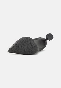 Zapato negro de punta afilada con un material superior suave, diseño elegante, suela de goma texturizada con marca impresa y una pequeña lengüeta en la parte trasera.