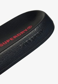 Tongs en caoutchouc noir avec surface texturée et logo "SUPERDRY" en relief de couleur rouge. Conçues avec une forme incurvée pour le confort et l'adhérence.
