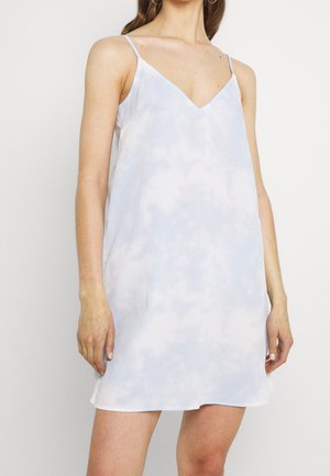 Kobieta w krótkiej, jasnoniebieskiej sukience typu slip w stylu tie-dye z cienkimi ramiączkami spaghetti i dekoltem w kształcie litery V, stojąca na jednolitym białym tle.