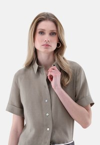 Camicia a maniche corte con colletto in tessuto marrone chiaro, caratterizzata da bottoni sul davanti e una vestibilità relax. Texture liscia, design minimale.
