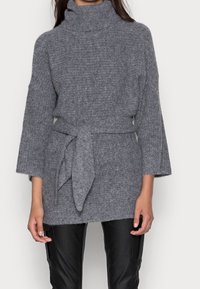 Grijze gebreide trui met een hoge hals, een ceintuur in de taille en brede mouwen, met een zachte textuur en een relaxte fit. Gepaard met zwarte broek.