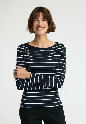 Donna sorridente con capelli castani corti che indossa una maglia nera a maniche lunghe con righe orizzontali bianche, in piedi con le braccia incrociate.