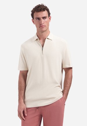 Mann trägt ein cremefarbenes, kurzärmeliges Polo mit Reißverschluss am Kragen und pinke Hose, steht mit einer Hand in der Tasche vor weißem Hintergrund.