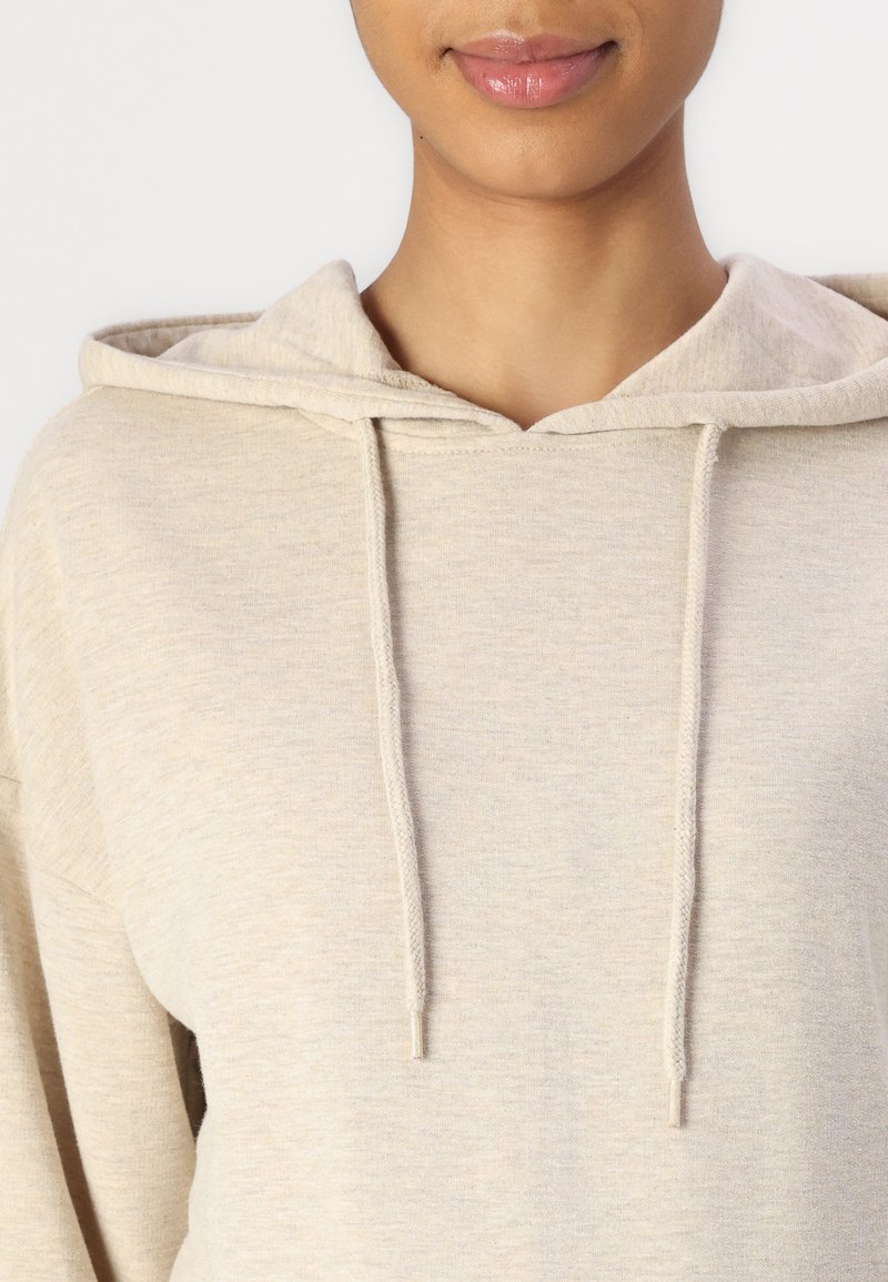 Helle beige Kapuzenjacke aus einem weichen, strukturierten Stoff, mit einer Kordelzugkapuze und einem lässigen Schnitt. Sichtbare Nähte und Hardware-Akzente.